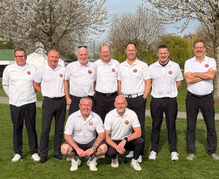 0e36bd84-719b-4d92-97d7-f1eab553a045 Eine Gruppe von neun Männern in weißen Polohemden steht und kniet auf einer Wiese vor Bäumen, alle lächeln in die Kamera.