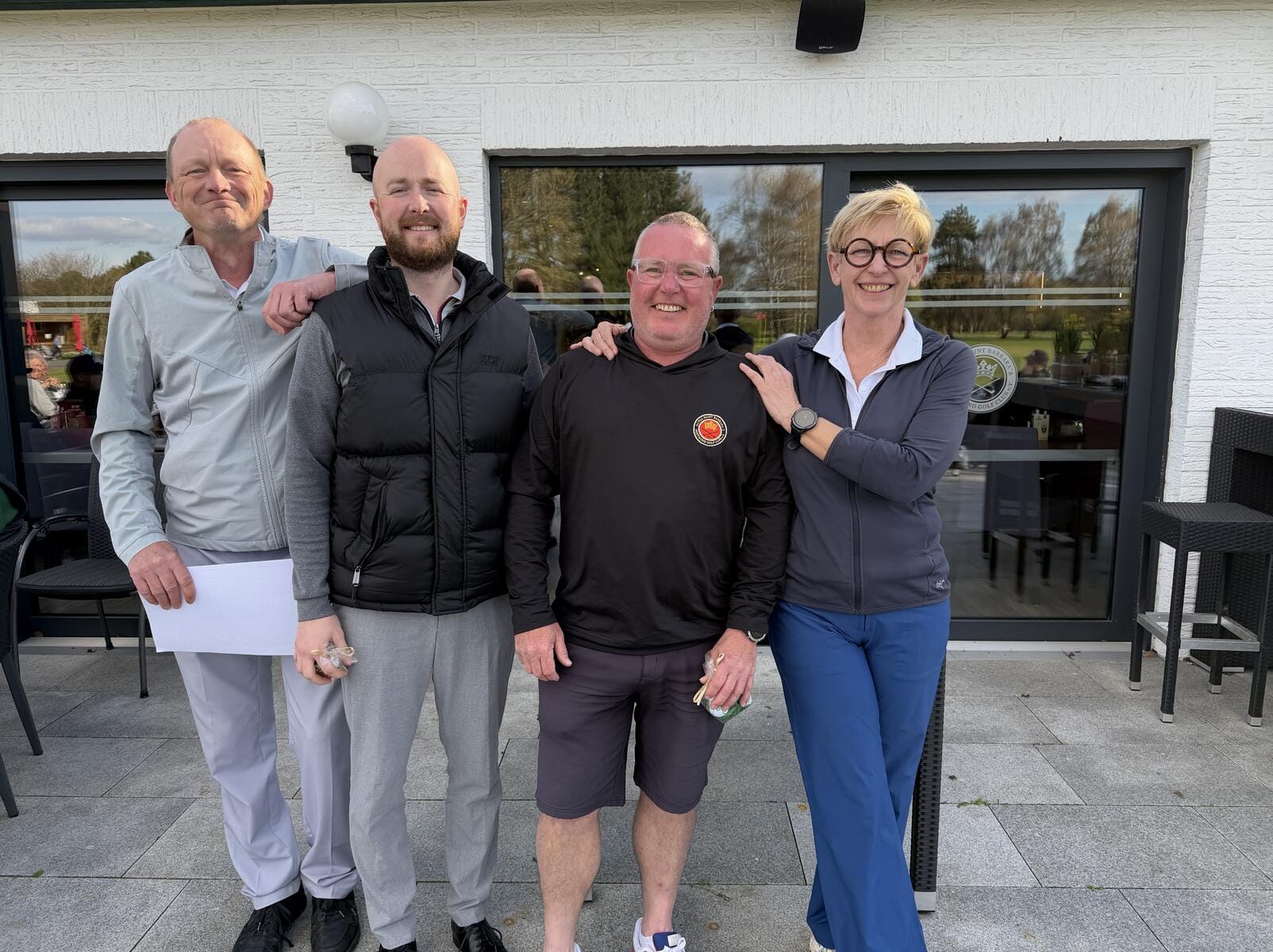 Vier Menschen stehen nebeneinander vor einem Gebäude mit großen Fenstern, lachen und wirken fröhlich, die Frau hat den Arm um die drei Männer.