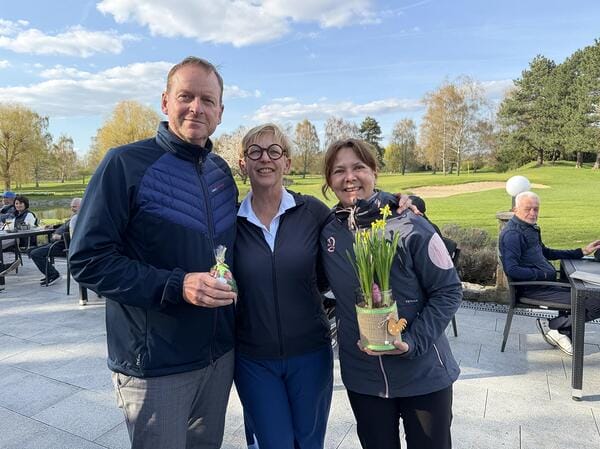 Drei fröhliche Menschen mit Blumen- und Schoko-Eiern in der Hand, draußen auf einer Terrasse bei schönem Wetter.
