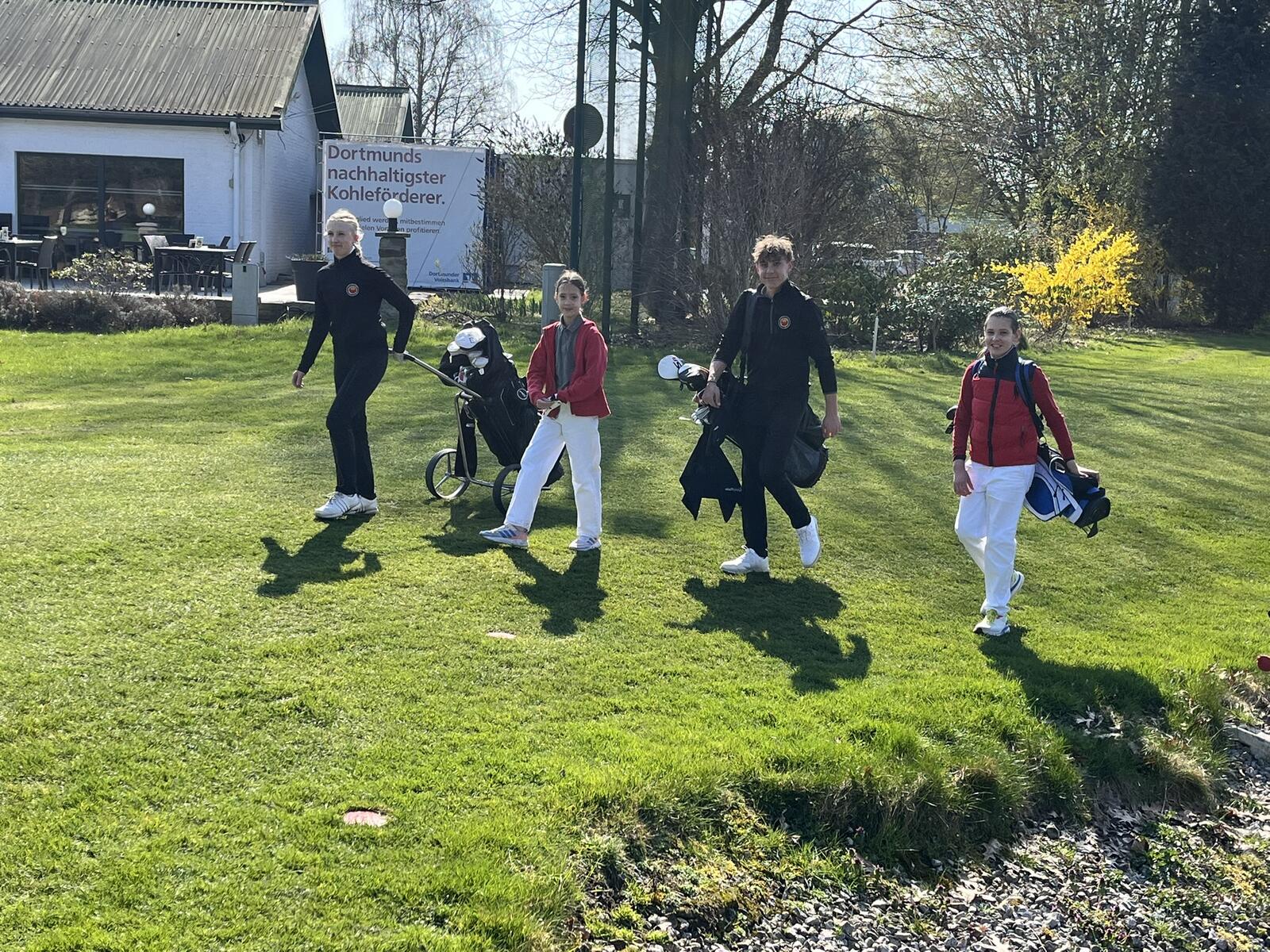 Vier junge Golferinnen gehen auf einem Rasen, einige mit Golfbags und -zubehör, in einer sonnigen Außenanlage.