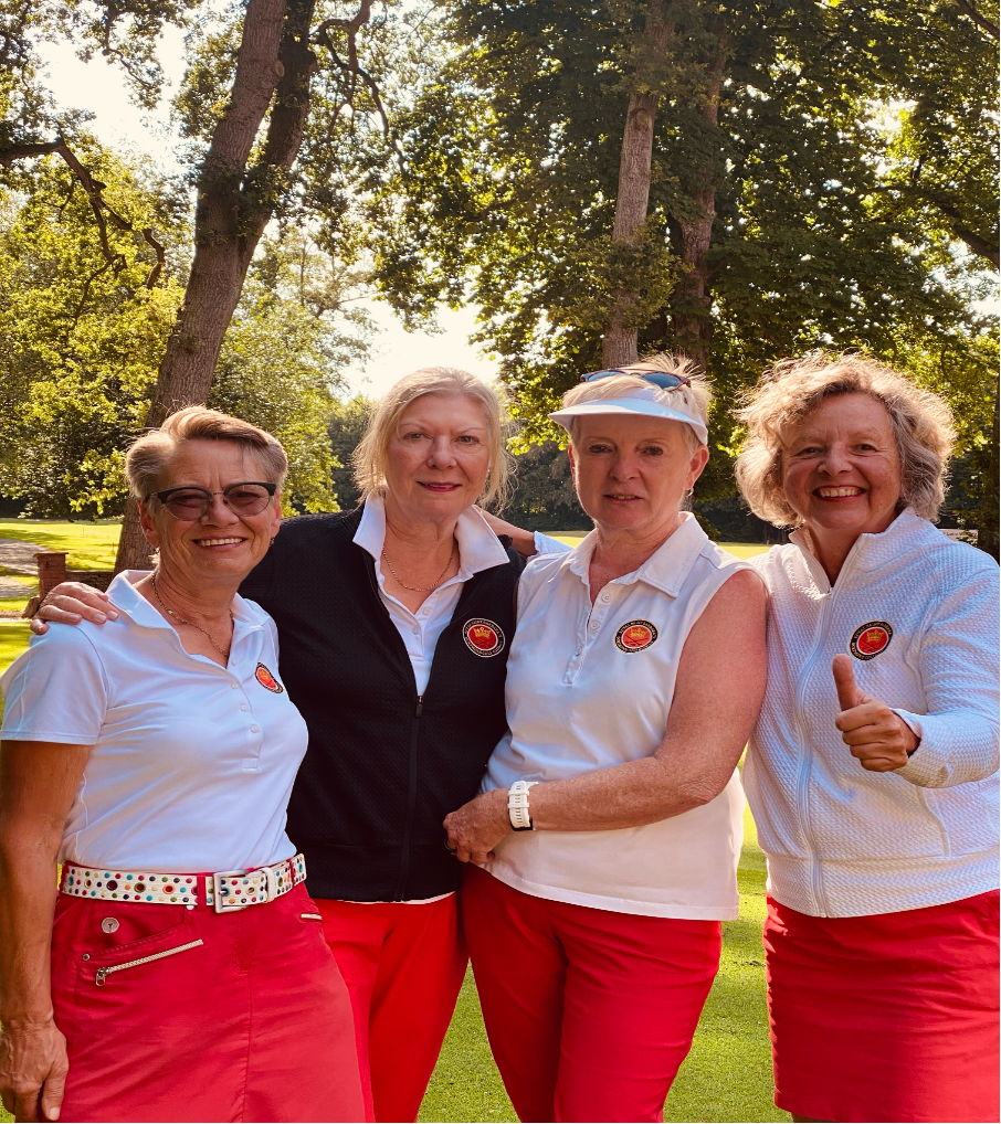 Vier ältere Frauen in Golfkleidung stehen im Freien auf einem Golfplatz, umgeben von Bäumen, sie lachen und posieren für die Kamera.