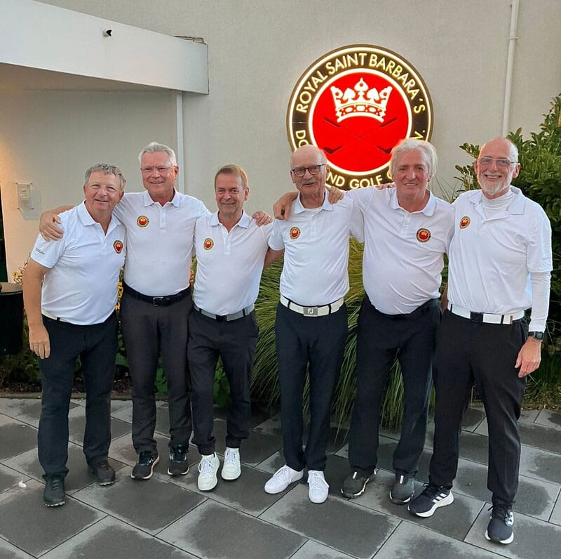 Sieben Männer in weißen Poloshirts mit einem Logo stehen nebeneinander vor einem Gebäude, auf dem ein leuchtendes Schild mit der Aufschrift "Royal Saint Barbara's" und einem roten Kronen- und Golfsymbol zu sehen ist.