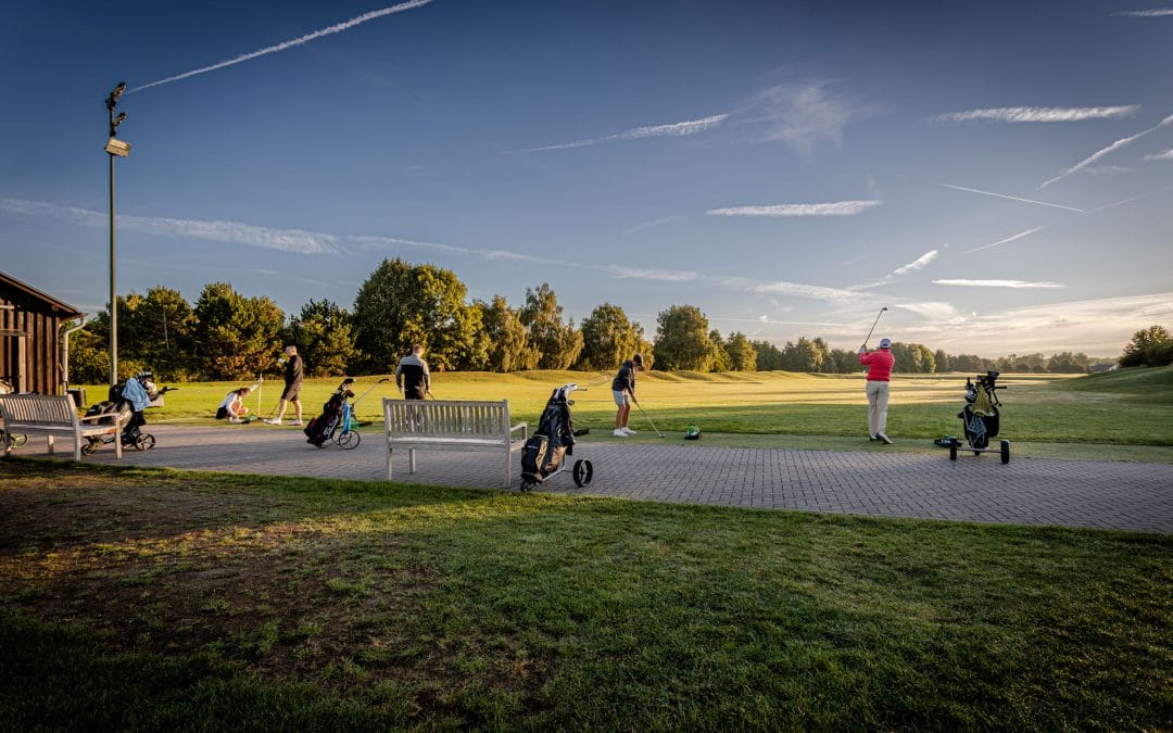 Mehrere Menschen beim Golfspielen auf einem Grün bei Sonnenuntergang, mit Golftaschen und Schlägern, auf einer gepflegten Golfanlage mit Bäumen im Hintergrund.