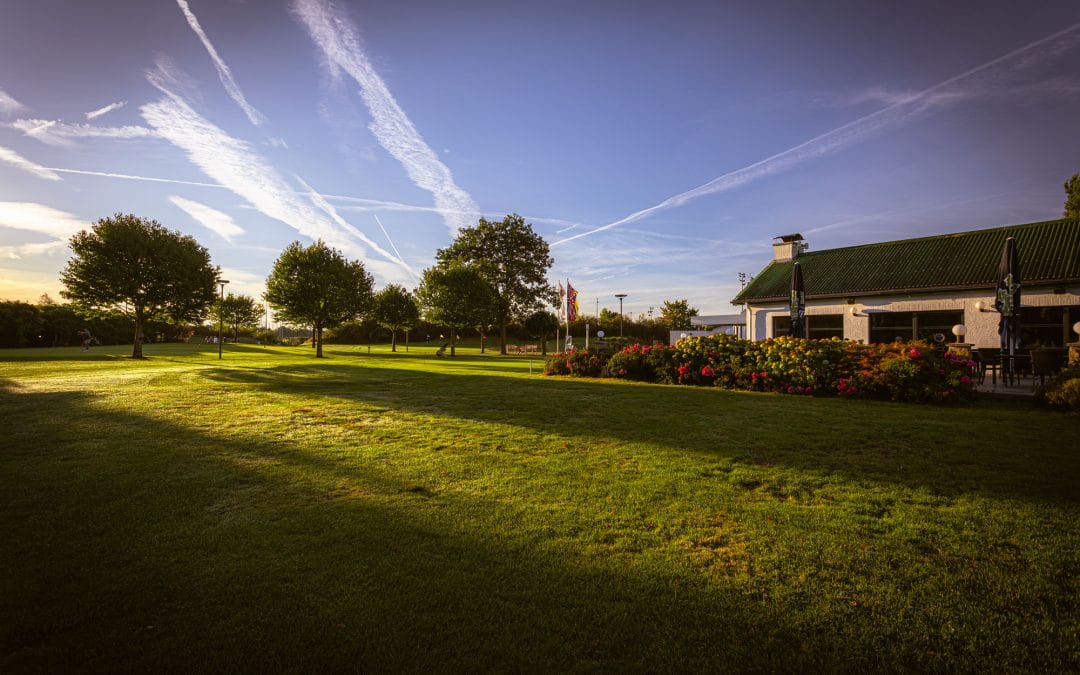 Ein sonniger Park mit großen Bäumen, einem gepflegten Grünflächenbereich und einem angrenzenden Gebäude mit Terrassen, auf denen Tische mit Sonnenschirmen stehen, bei klarem Himmel mit sichtbaren Kondensstreifen.