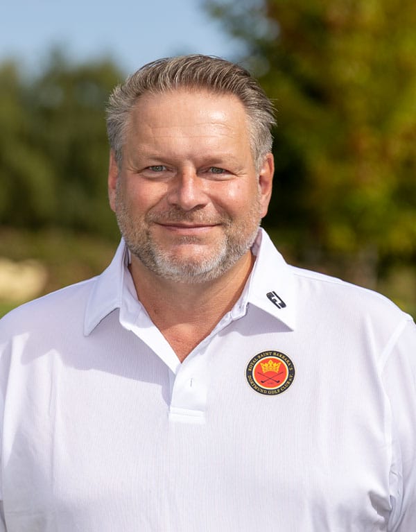 Ein Mann mit kurzgrauem Haar und Bart, trägt ein weißes Polo-Shirt mit einem Logo auf der linken Brust, steht im Freien mit unscharfem, herbstlichem Hintergrund.