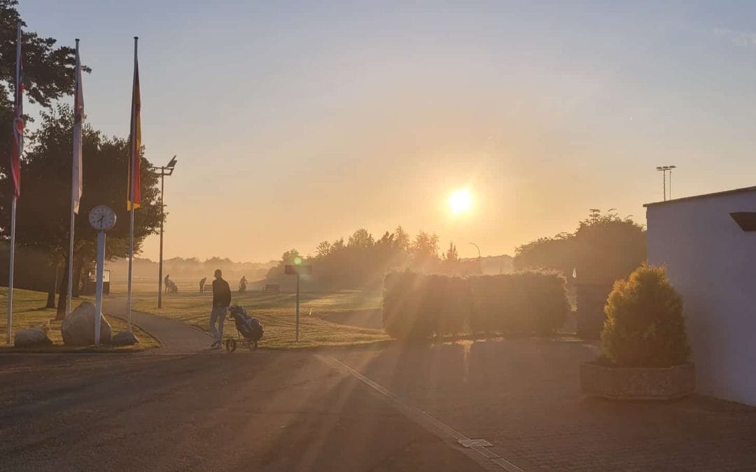 Sonnenaufgang über einem Golfplatz mit Personen, die Golf spielen, Bäumen, Gebäuden und Flaggen in der Morgendämmerung