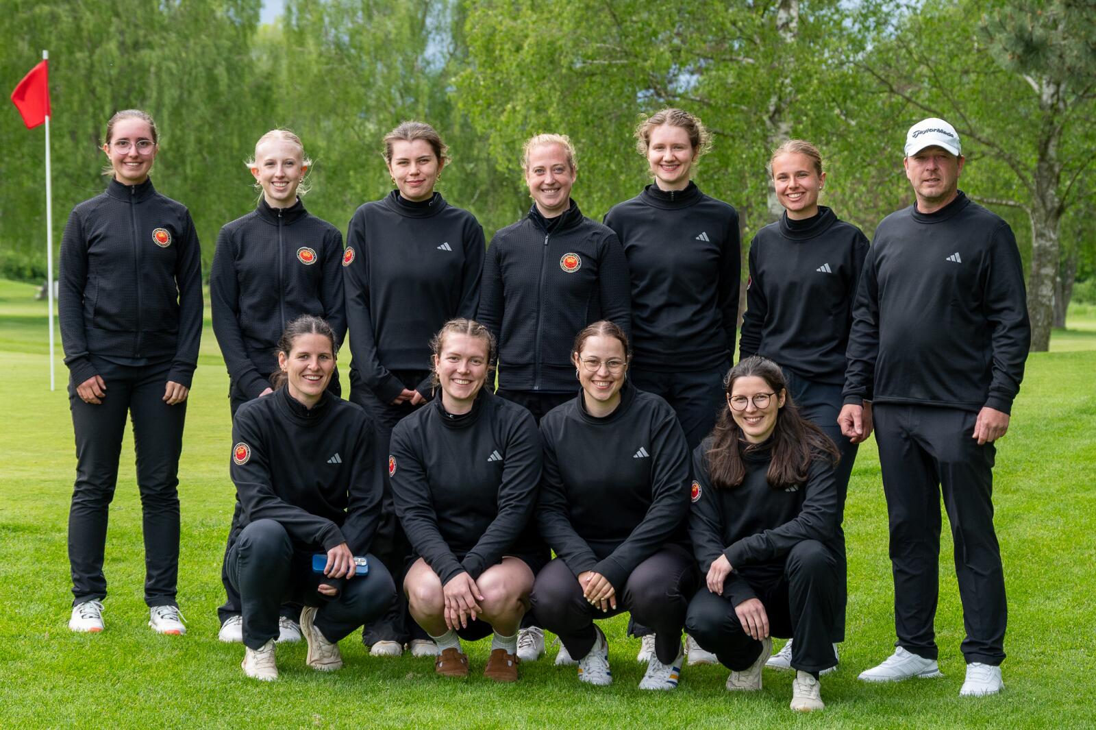 Eine Gruppe von elf Personen, hauptsächlich Frauen, steht und kniet auf einem grünen Golfplatz bei schönem Wetter.
