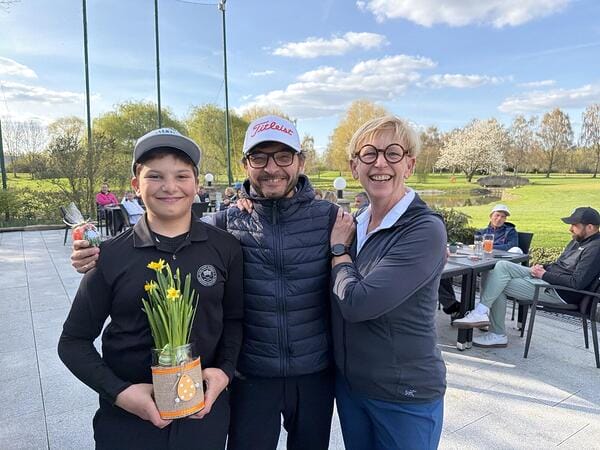 Drei Menschen stehen zusammen outdoors, lachen und halten einen Topfpflanzen mit gelben Narzissen. Es ist sonnig, und im Hintergrund sind weitere Personen, Bäume und ein Teich sichtbar.
