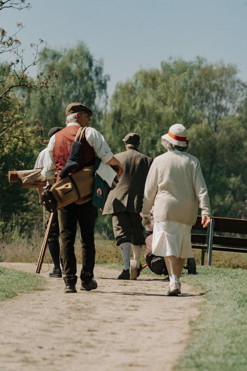Ältere Menschen gehen gemeinsam im Park spazieren, umgeben von Bäumen bei schönem Wetter.