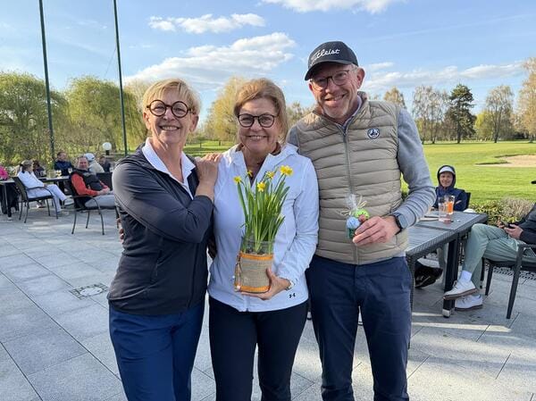 Drei lachende Personen stehen im Freien, eine hält eine Vase mit gelben Blumen, im Hintergrund sitzen weitere Menschen auf Terrassenmöbeln.