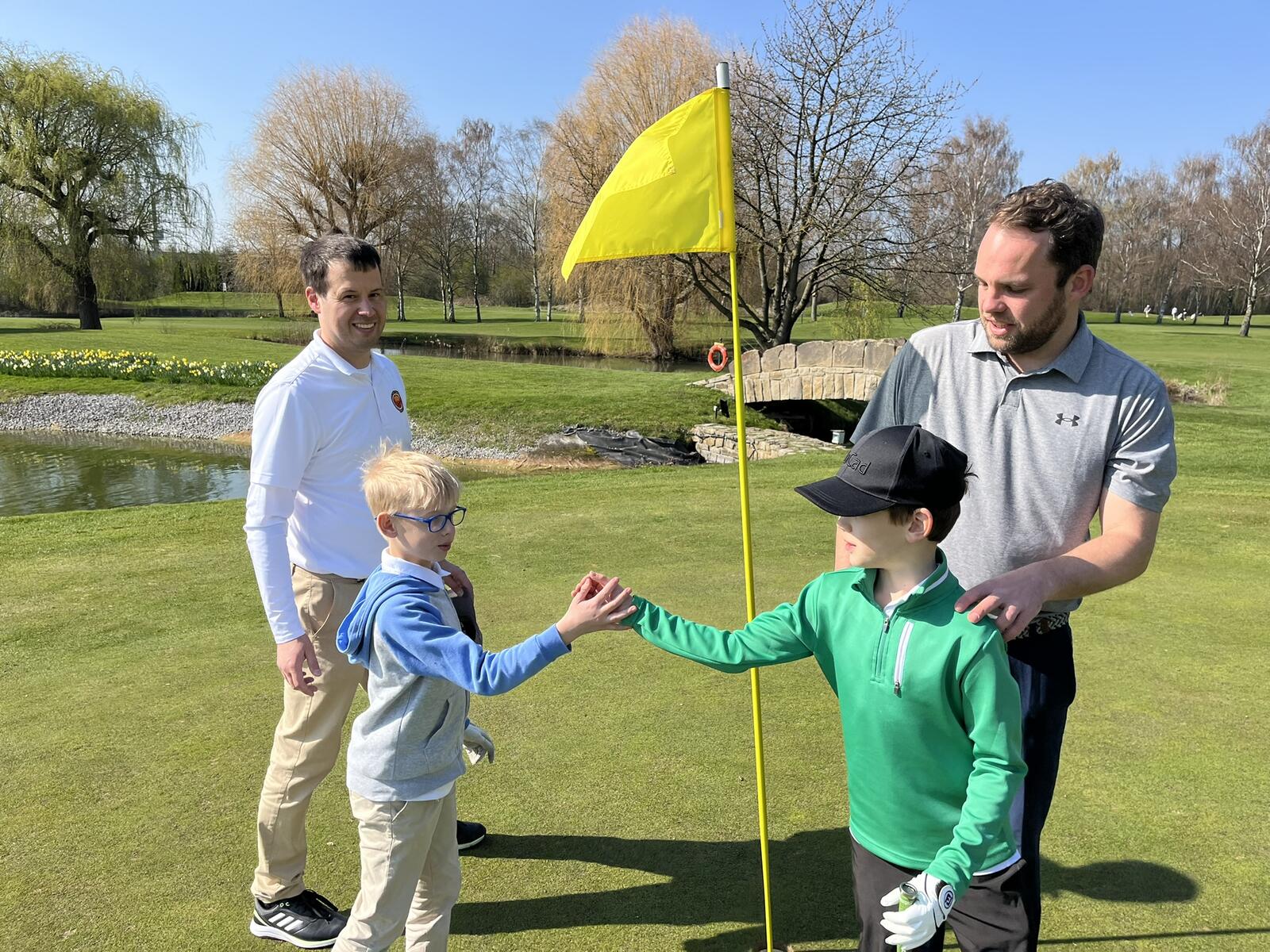Zwei Jungen, die sich die Hand geben, stehen an einem Golfplatz neben einer Flagge, während zwei Männer im Hintergrund lächelnd zuschauen.