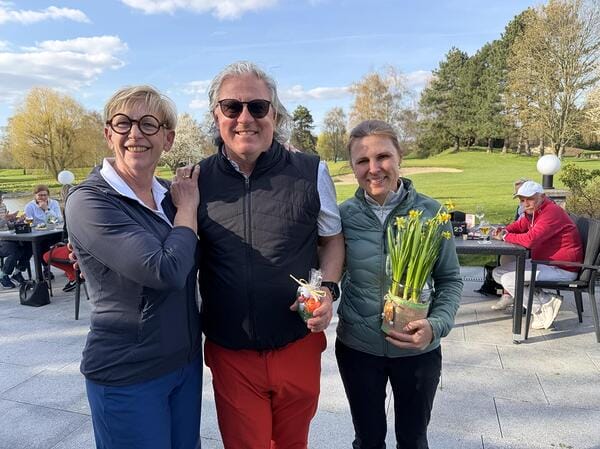 Drei Personen stehen zusammen outdoors vor einer grünen Golfanlage, die mittlere Person trägt dunkle Sonnenbrille, hält eine kleine Geschenkverpackung, während die rechte Person einen Topf mit gelben Blumen hält, alle lächeln.
