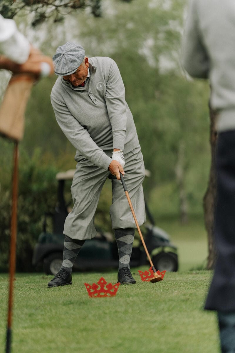 Ein Mann in Golfkleidung schlägt den Ball auf dem Grün, trägt einen grauen Pullover, Hut und argyle-Socken.