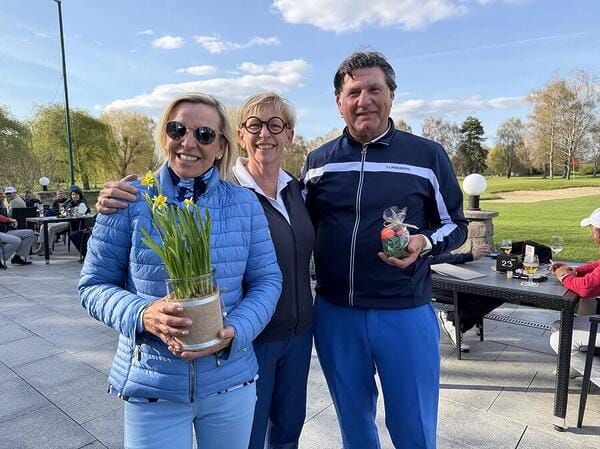 Drei Menschen stehen auf einer Terrasse im Freien, lachen und halten Blumen und ein Geschenk, im Hintergrund sind weitere Personen beim Golfen und Essen.
