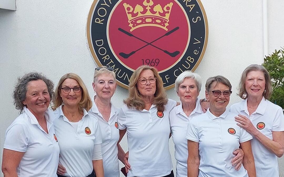 Sieben Frauen in weißen Polo-Shirts stehen nebeneinander vor einem weißen Wand mit einem großen schwarzen und roten Vereinslogo, das einen goldenen Krone, zwei Schläger und das Jahr 1969 zeigt.