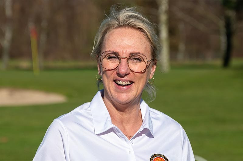 Eine lachende Frau mit Kurzhaarschnitt, Brille und Ohrringen steht im Freien auf einer Wiese mit Bäumen im Hintergrund.