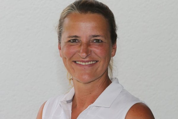 Eine lachende Frau mit braunen Haaren in einem weißen Sporttop vor glänzendem, hellgrauem Hintergrund.