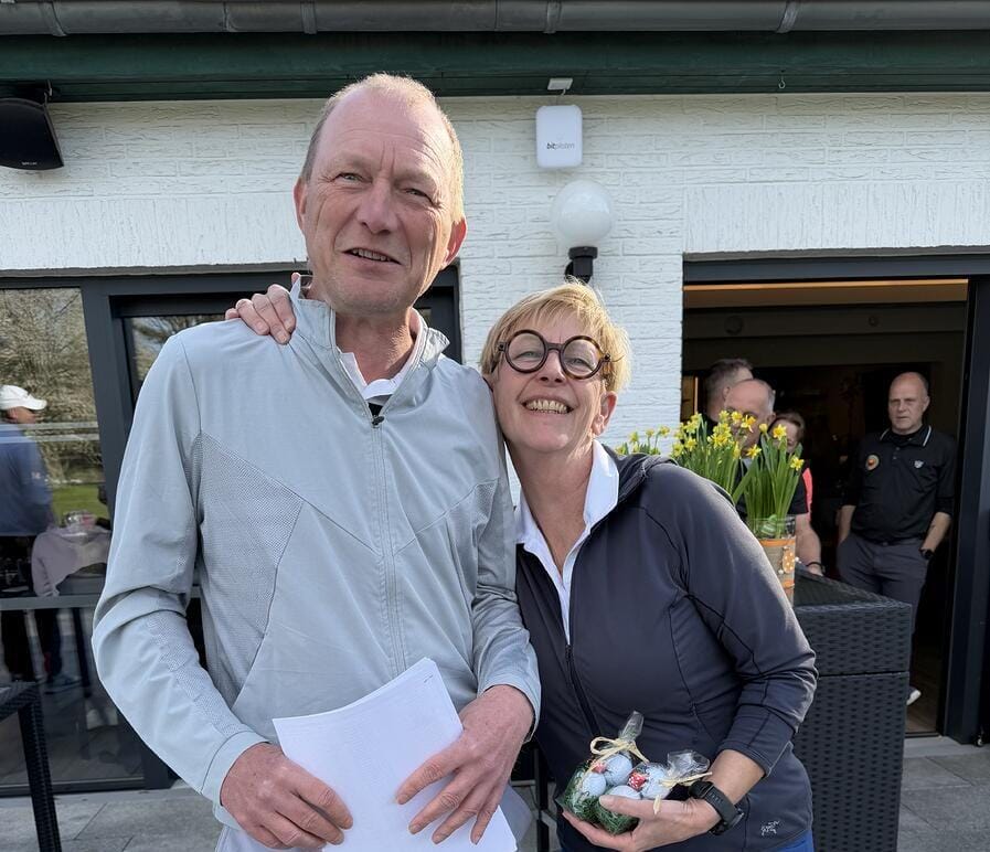 A man and a woman are smiling and standing close together outdoors in front of a building, with the woman holding a small gift bag.