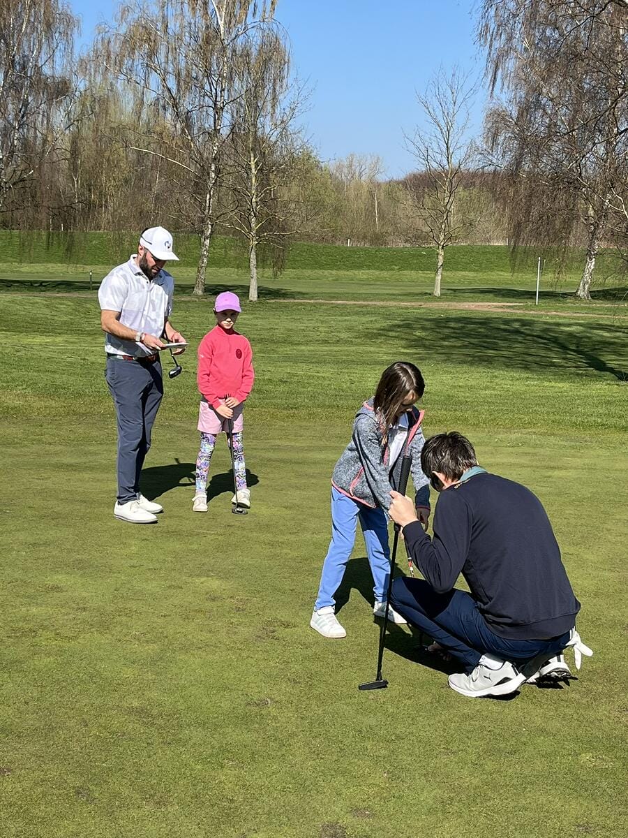 Ein Mann zeigt einer jungen Mädchen auf einem Golfplatz, wie man einen Golfschläger benutzt, während zwei weitere Kinder zuschauen.