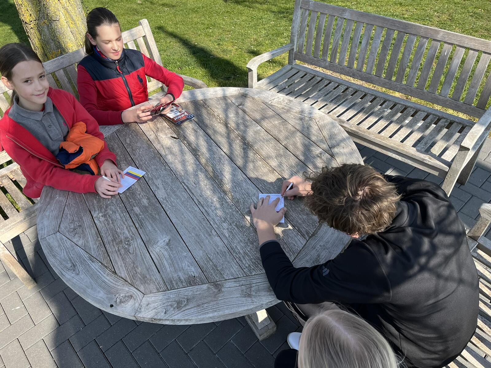 Drei Kinder sitzen am Tisch mit einem Mann, spielen Karten, während die Sonne scheint.