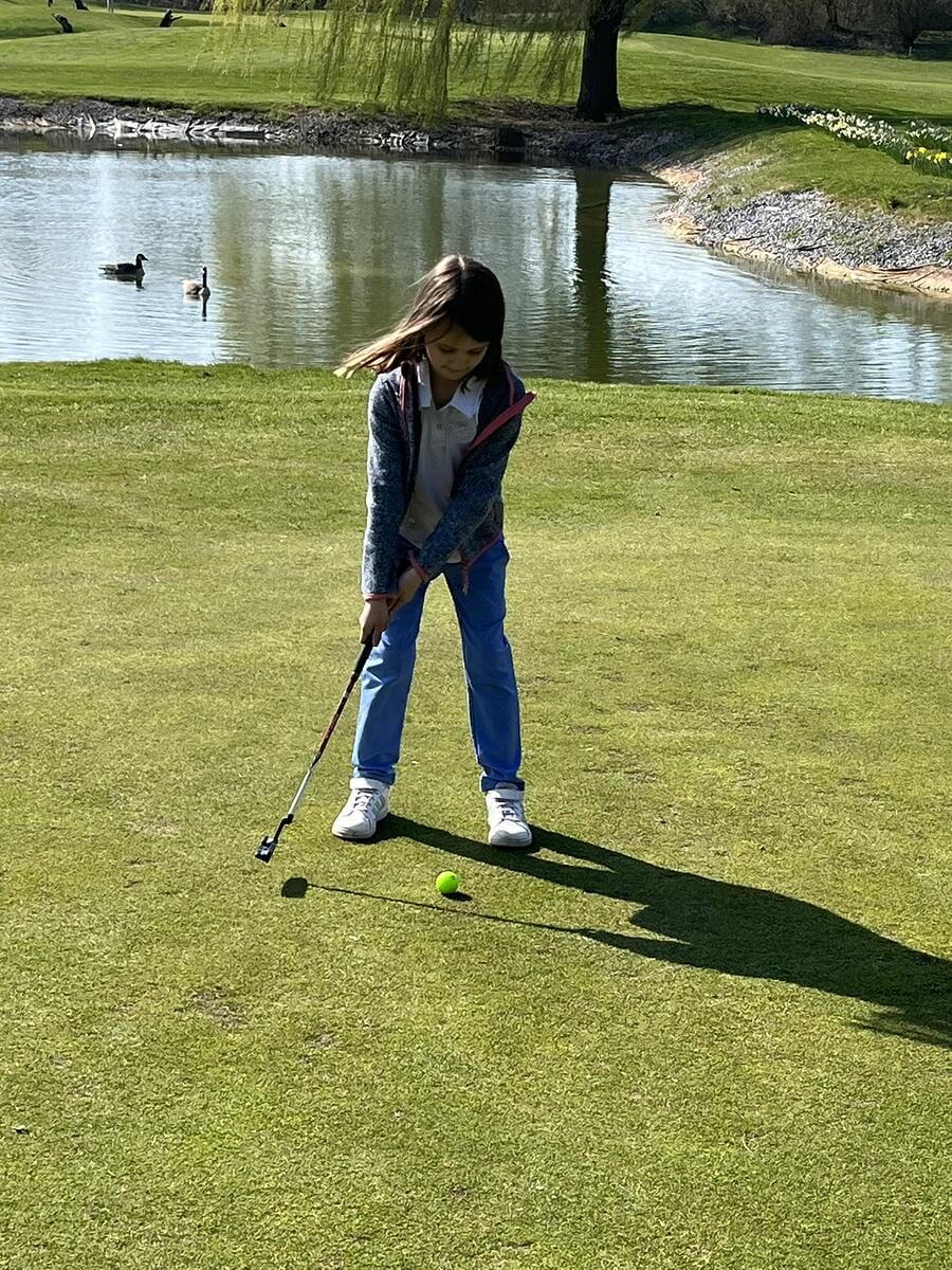 Ein Mädchen beim Golfspielen auf einem Grün, mit einem See und Enten im Hintergrund.
