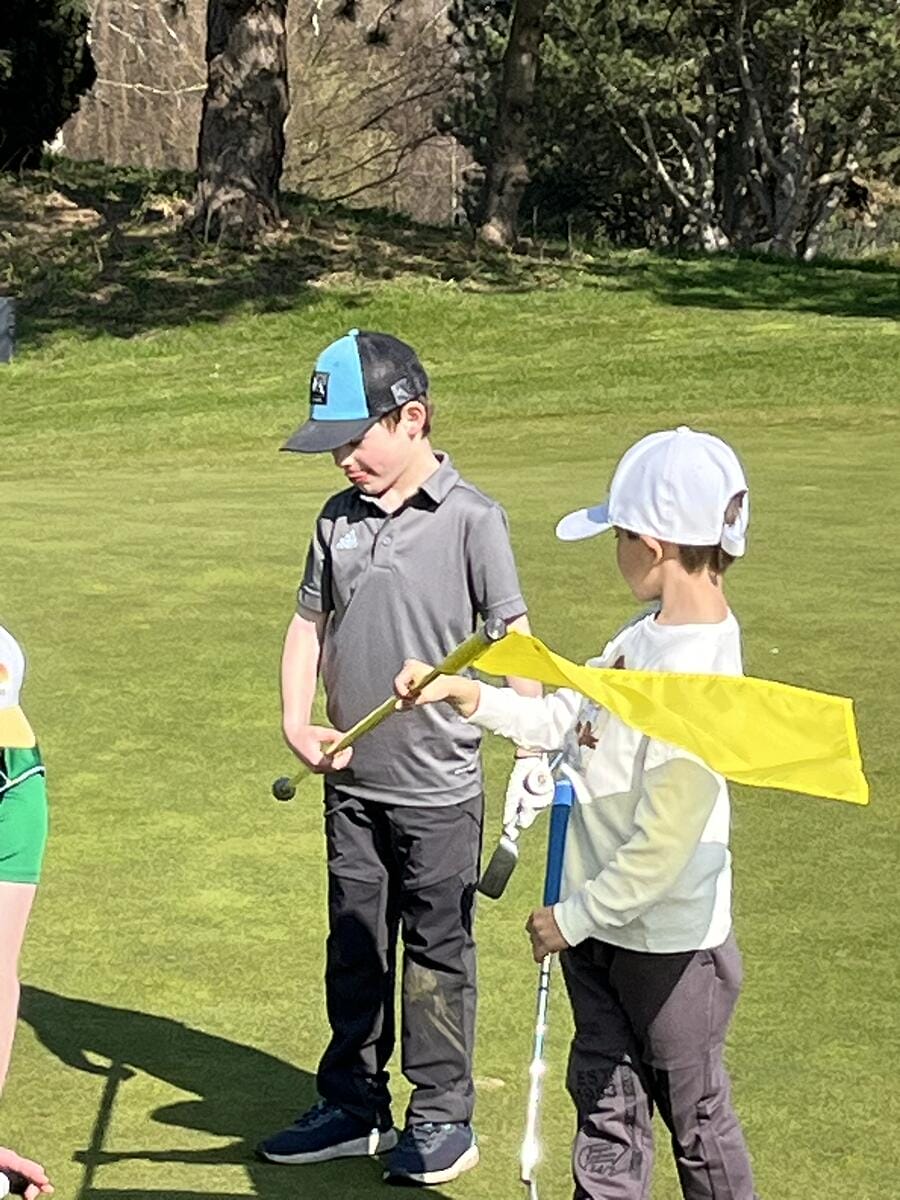 Zwei Jungen auf einem Golfplatz, einer hält eine Flagge und der andere einen Schläger, bei sonnigem Wetter.