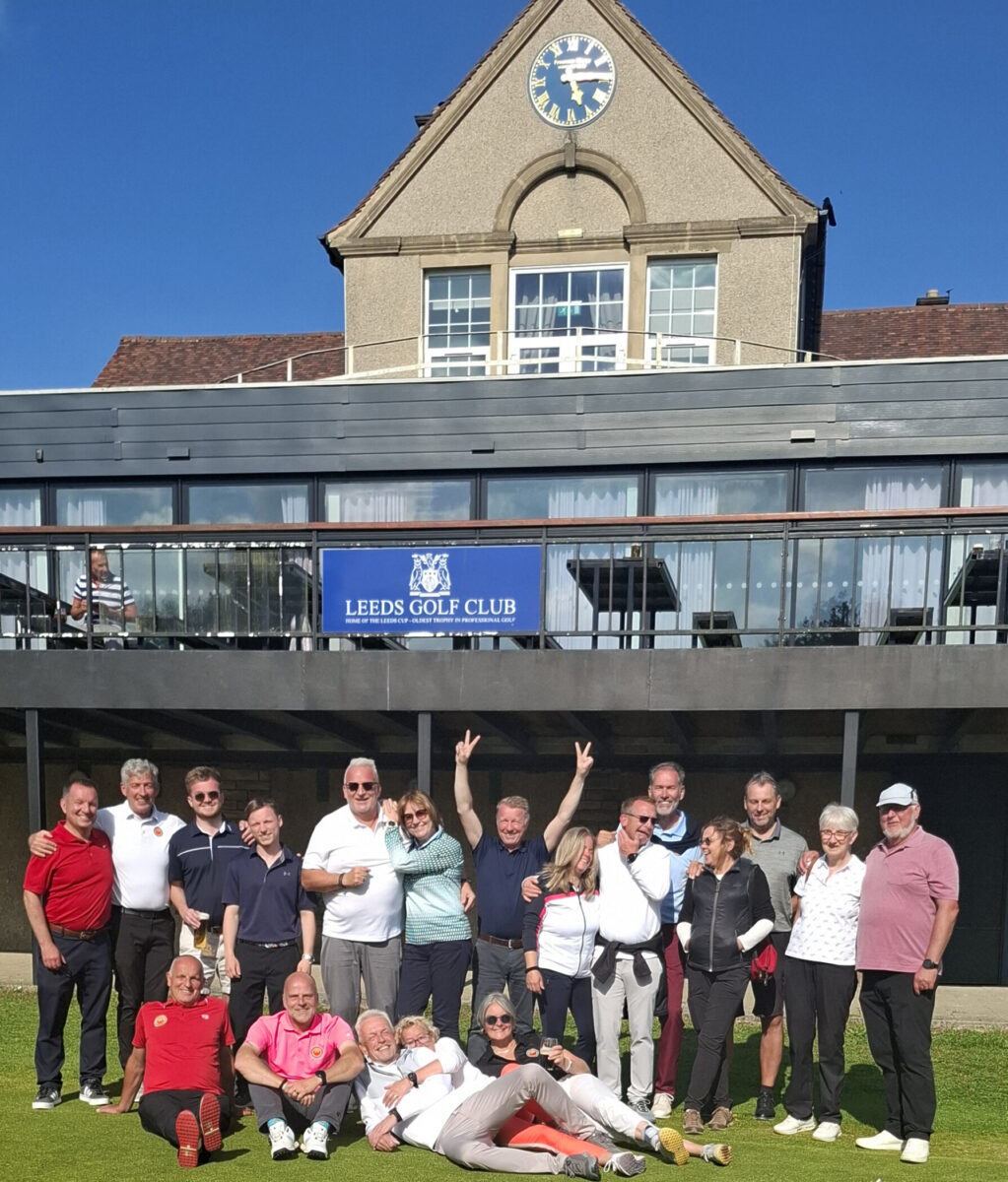 Gruppenfoto von mehreren Menschen, die auf dem Grass ansehen, vor einem Gebäude mit einer signierten Bank, oberhalb eines Clubschildes, im Freien bei sonnigem Wetter.
