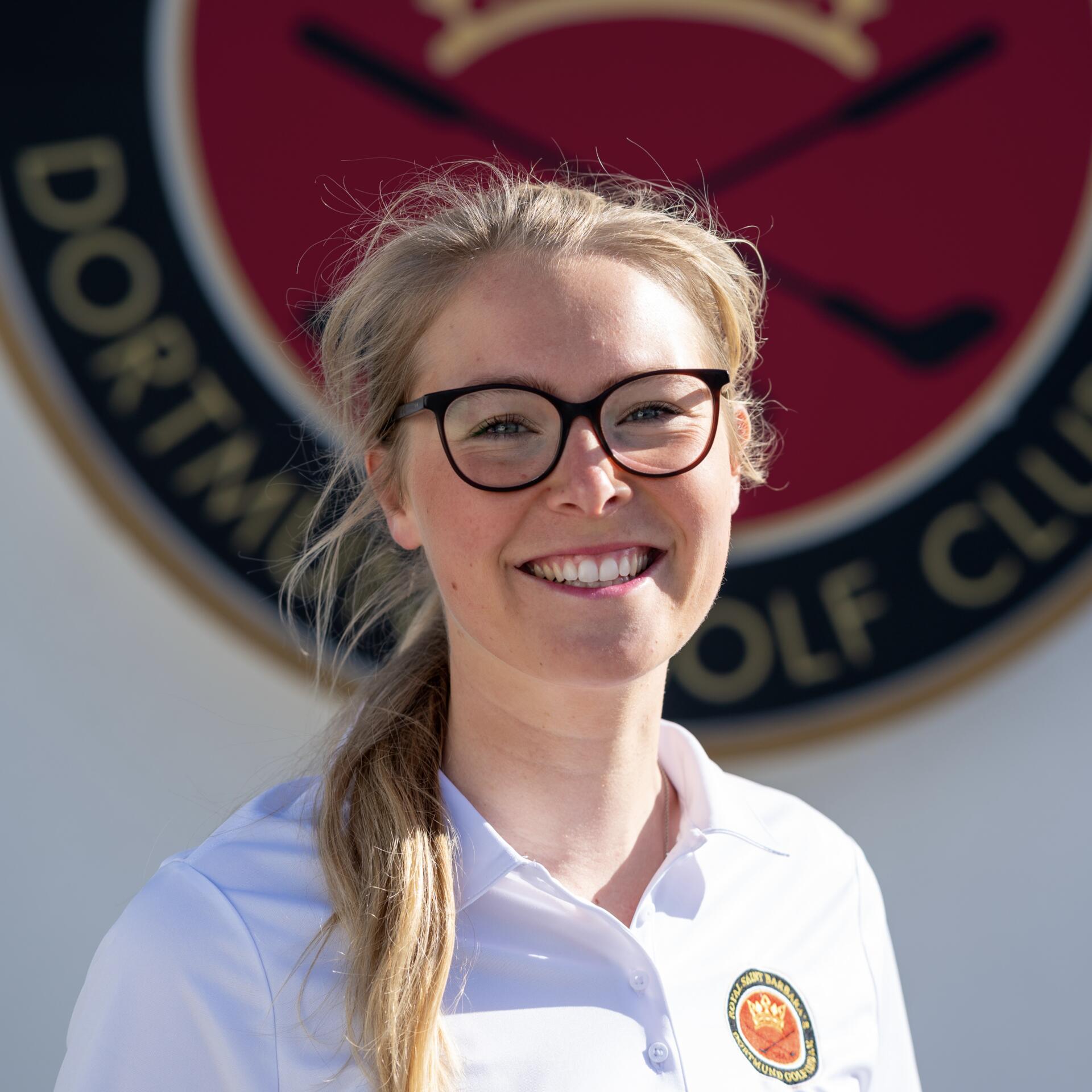 Junge Frau mit blonden Haaren und sommerlichem Gesichtsausdruck, draußen auf einer Wiese, trägt eine dunkelblaue Polo-Bluse und hält einen Sport-Schläger über die Schulter.
