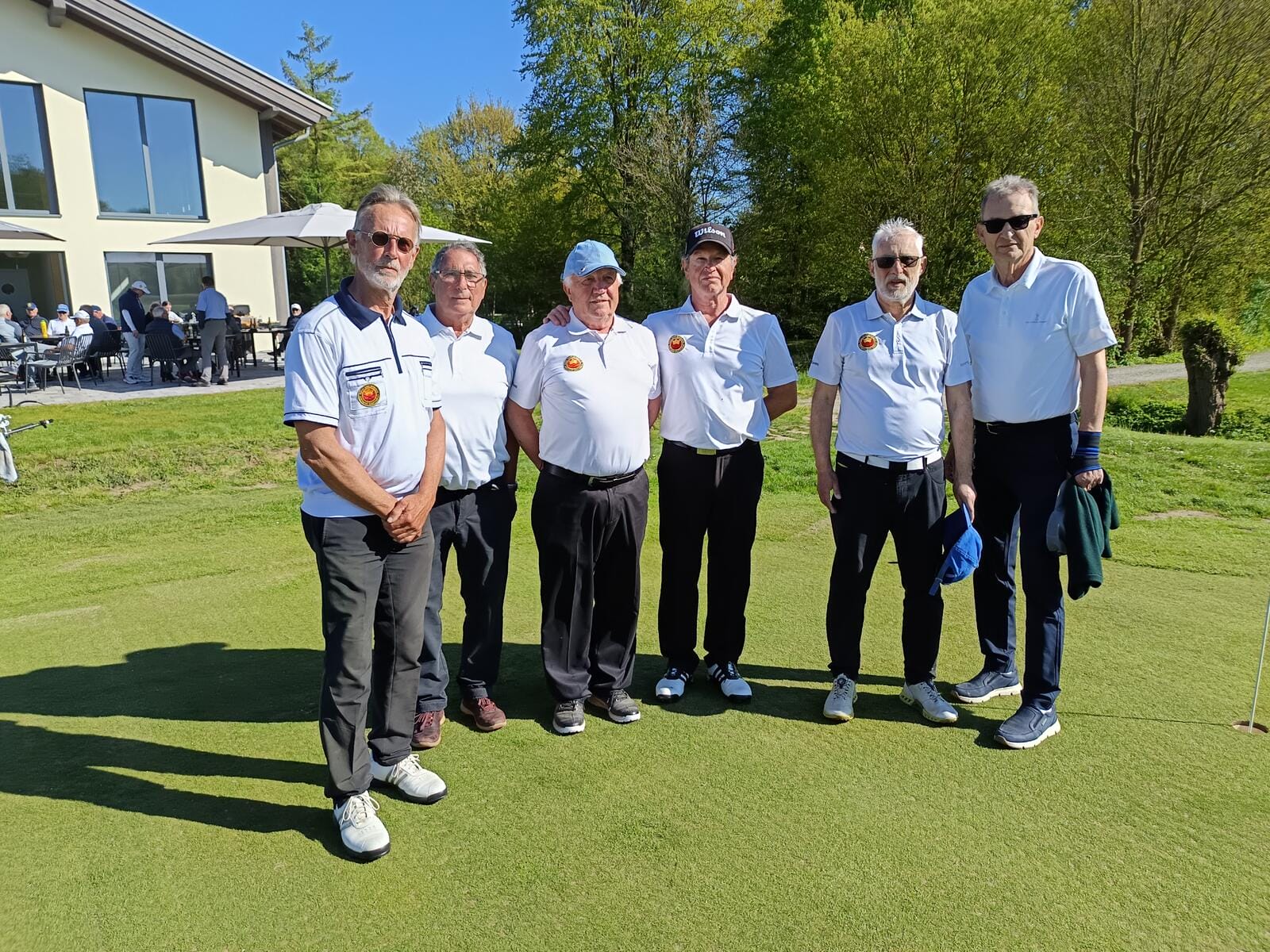 Sieben Männer stehen auf einem Golfplatz, alle tragen weiße Polohemden und schwarze Hosen, einige mit Kappen oder Sonnenbrillen, bei sonnigem Wetter.