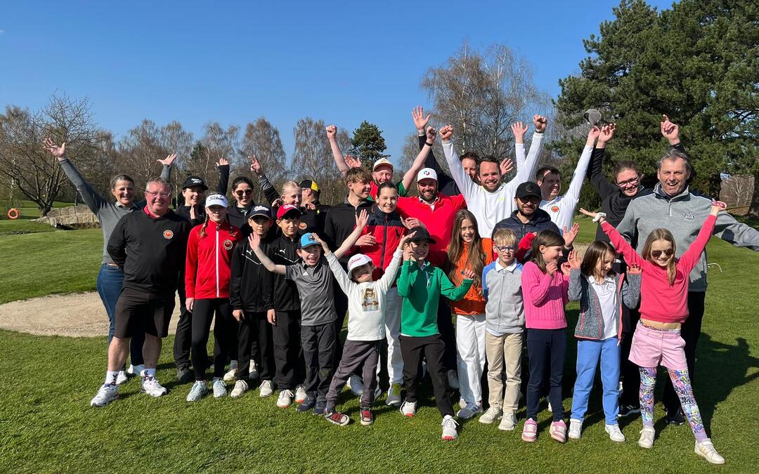 Eine Gruppe von Menschen, darunter Kinder und Erwachsene, freut sich fröhlich zusammen auf einem grünen Golfplatz bei strahlendem Sonnenschein.