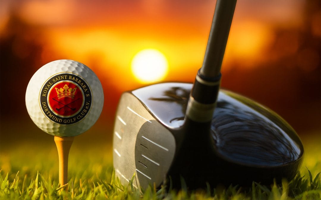 Ein Golfball mit einem Rotgold-Emblem der Royal Saint Barbara's Dortmund Golf Club auf einem Tee und ein Golfschläger auf dem Rasen vor einem Sonnenuntergang im Hintergrund.