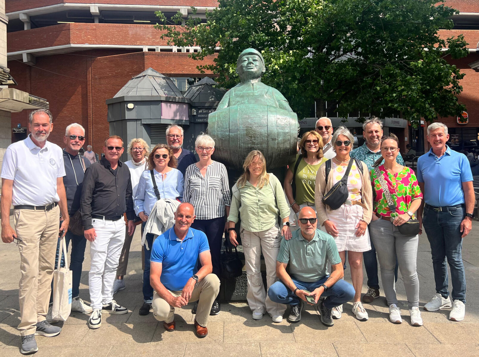 Eine Gruppe von 17 Erwachsenen steht vor einem großen, grünen Skulptur-Depot in einer Stadt, im Hintergrund sind rote Backsteingebäude und eine große grüne Baum.