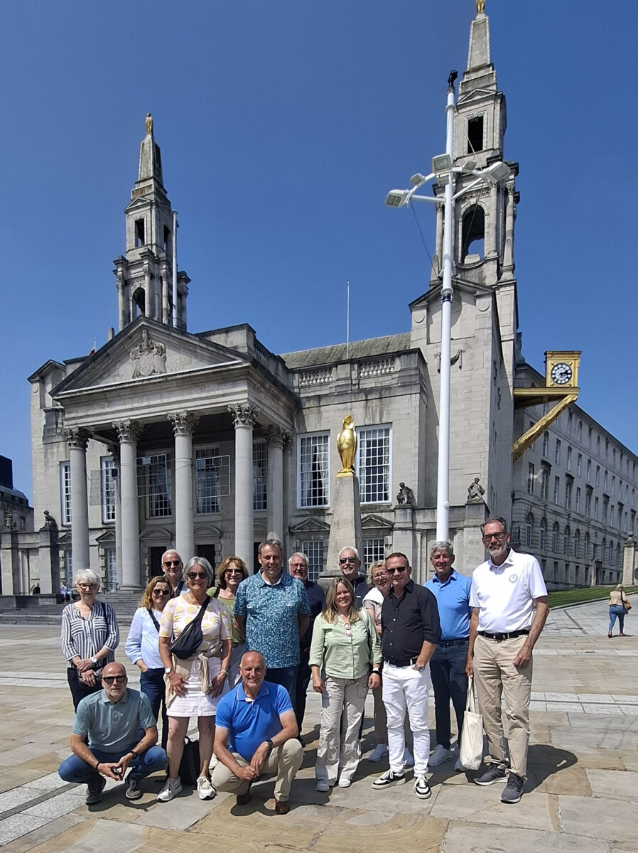 Eine Gruppe von Menschen steht vor einer großen, historischen Kathedrale mit zwei hohen Türmen und goldenen Verzierungen, bei strahlendem Sonnenschein und klarem blauen Himmel.