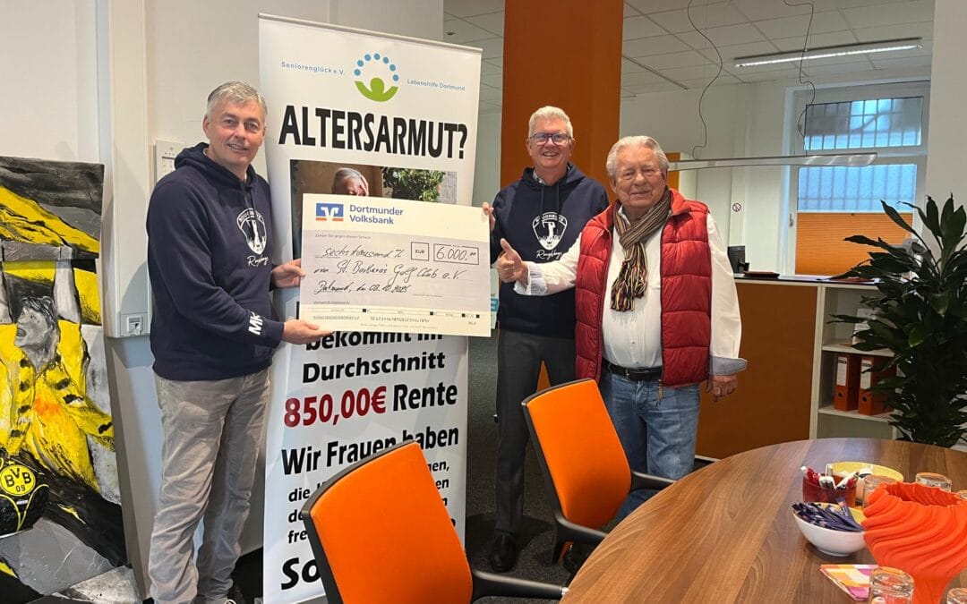 Drei Männer stehen in einem Raum, der einen großen Scheck über 6000 Euro halten, während sie vor einem Banner stehen, das eine Diskussion über Armut im Alter beschreibt, mit einer farbigen Wand im Hintergrund und einem Raum mit Tischen und Stühlen.