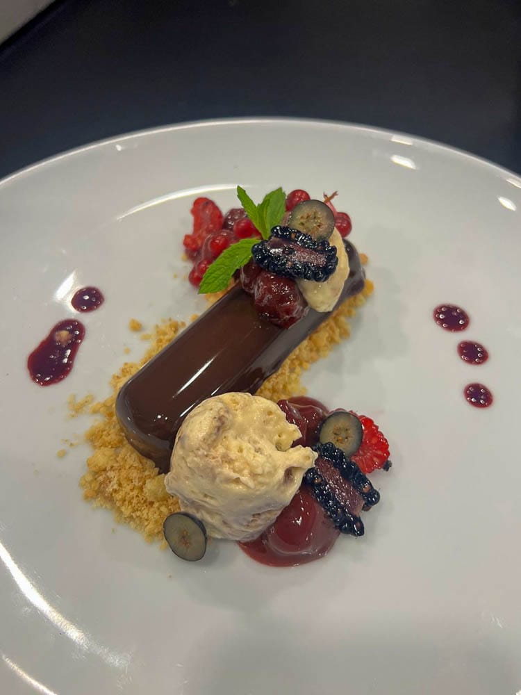 Dessert mit einem rechteckigen Schokoladenkuchen, einer Kugel Vanilleeis, Beeren, Minzblatt, Früchte und einem essbaren Dekor auf einem weißen Teller