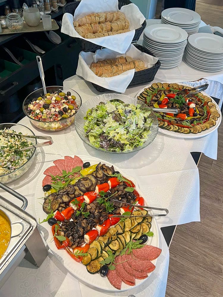 Ein Buffet mit verschiedenen Salaten, einer Platte mit gegrilltem Gemüse, Wurstwaren und frischem Brot, alles auf einem weißen Tischtuch angerichtet.