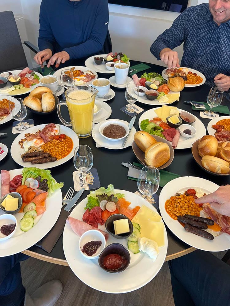 Tisch mit mehreren Tellern voller Frühstück, darunter verschiedene Wurst- und Fleischsorten, Käse, Brot, Lachs, Salat, Marmelade, Butter, Eier, Orangensaft und verschiedene Beilagen. Menschen sitzen um den Tisch, essen und haben Gläser und Tassen vor sich.