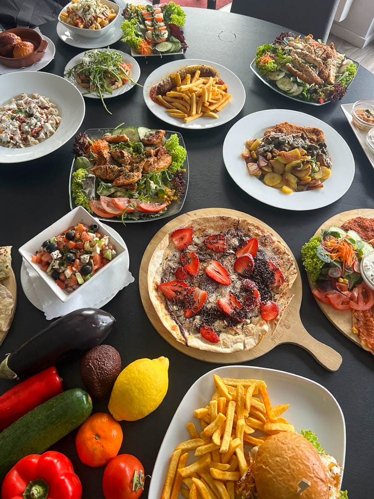 Teller mit verschiedenen italienischen Gerichten, darunter Pizza mit Erdbeeren, Pasta, Fleischgerichte, Salate, Gemüse und Pommes Frites, auf einem schwarzen Esstisch