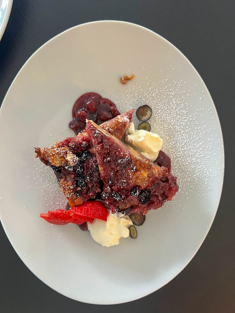 Ein Teller mit einem Stück Obstkuchen, garniert mit Beeren, einer Erdbeerscheibe, einer kleinen Kugel Eis, einem Klecks Sahne und Beeren, auf einem weißen Teller.