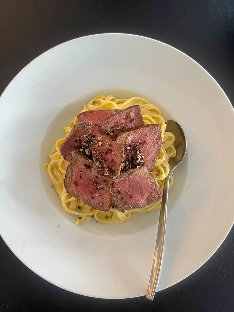 Ein weißer Teller mit Pasta und rosa gebratenem Fleisch, garniert mit schwarzen Pfefferkörnern, auf einem schwarzen Tisch, daneben ein Löffel.