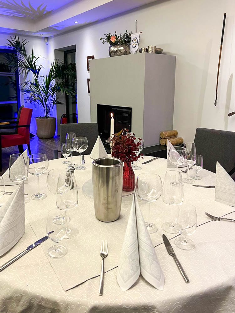 Ein festlich gedeckter runder Tisch mit weißer Tischdecke, mehreren Gläsern, Messer, Gabeln und gefalteten weißen Servietten, sowie einer roten Vase mit roten Blumen und einer Kerze in der Mitte. Im Hintergrund ist ein moderner Kamin, Dekorationen auf dem Kaminsims und eine große Zimmerpflanze. Es gibt auch einen dunklen Stab an der Wand links, eine rote Stuhl und einen großen Fensterbereich.