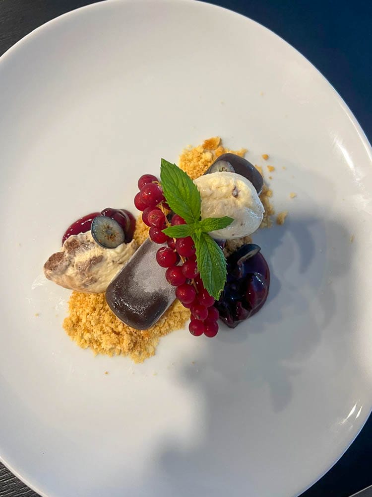 Ein Dessert mit einer Kugel Eis, umgeben von roten und blauen Beeren, Minzblättern, Keksbröseln und einem Schokoriegel auf einem weißen Teller.