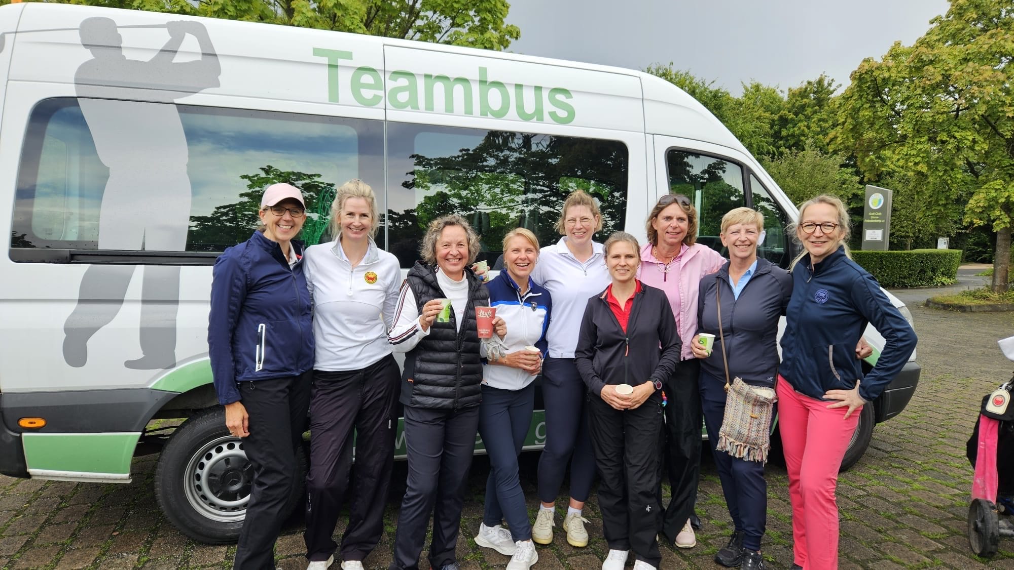 Gruppe von neun Frauen, die vor einem weißen Van mit grüner Beschriftung 'Teambus' stehen, lächelnd und mit Getränken in der Hand, in einer parkähnlichen Umgebung mit Bäumen und Gehwegen.