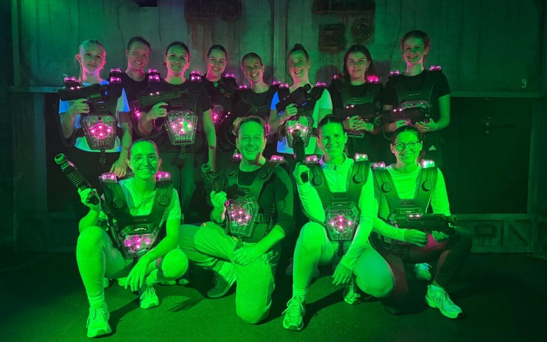 Eine Gruppe von elf Menschen in Neon-grünen und schwarzen Schutzwesten mit Laserwaffen, die in einem dunklen Raum mit grüner Beleuchtung für ein Laser-Tag-Spiel posieren.