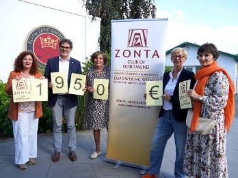 Fünf Personen halten die großen Check- und Preisschilder, während sie neben einem Zonta-Club-Stand mit einem Banner stehen.