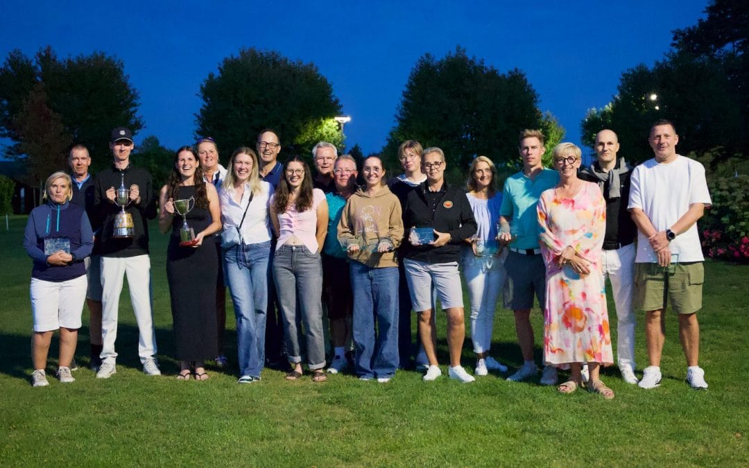 A group of people standing outdoors on a grassy area at night, holding trophies and awards, with trees and a darkening sky in the background.