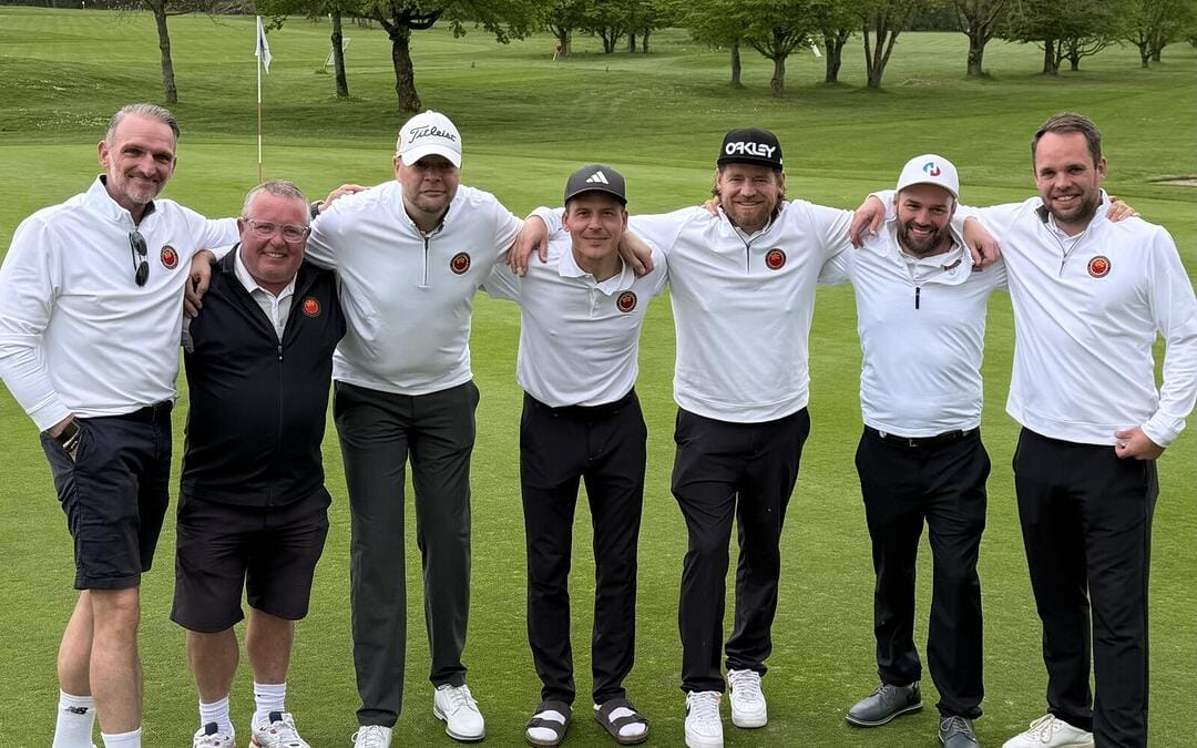 Sieben Männer stehen auf einem Golfplatz, Arm in Arm, lachend und in sportlicher Kleidung, bereit für ein Gruppenfoto.