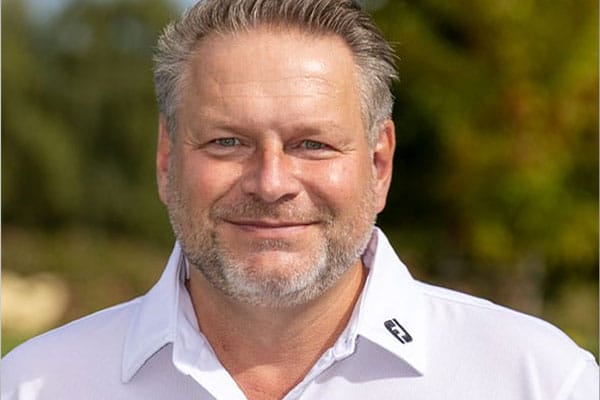 Ein lachender Mann mit grauem Haar und Bart, trägt ein weißes Hemd, im Hintergrund sind unscharfe grüne und braune Blätter.