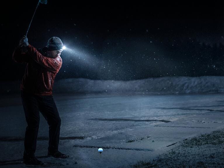 Ein Golfer schlägt bei Nacht auf einem verschneiten Platz mit einer Taschenlampe auf seinem Kopf; der Golfball ist auf dem Boden sichtbar.